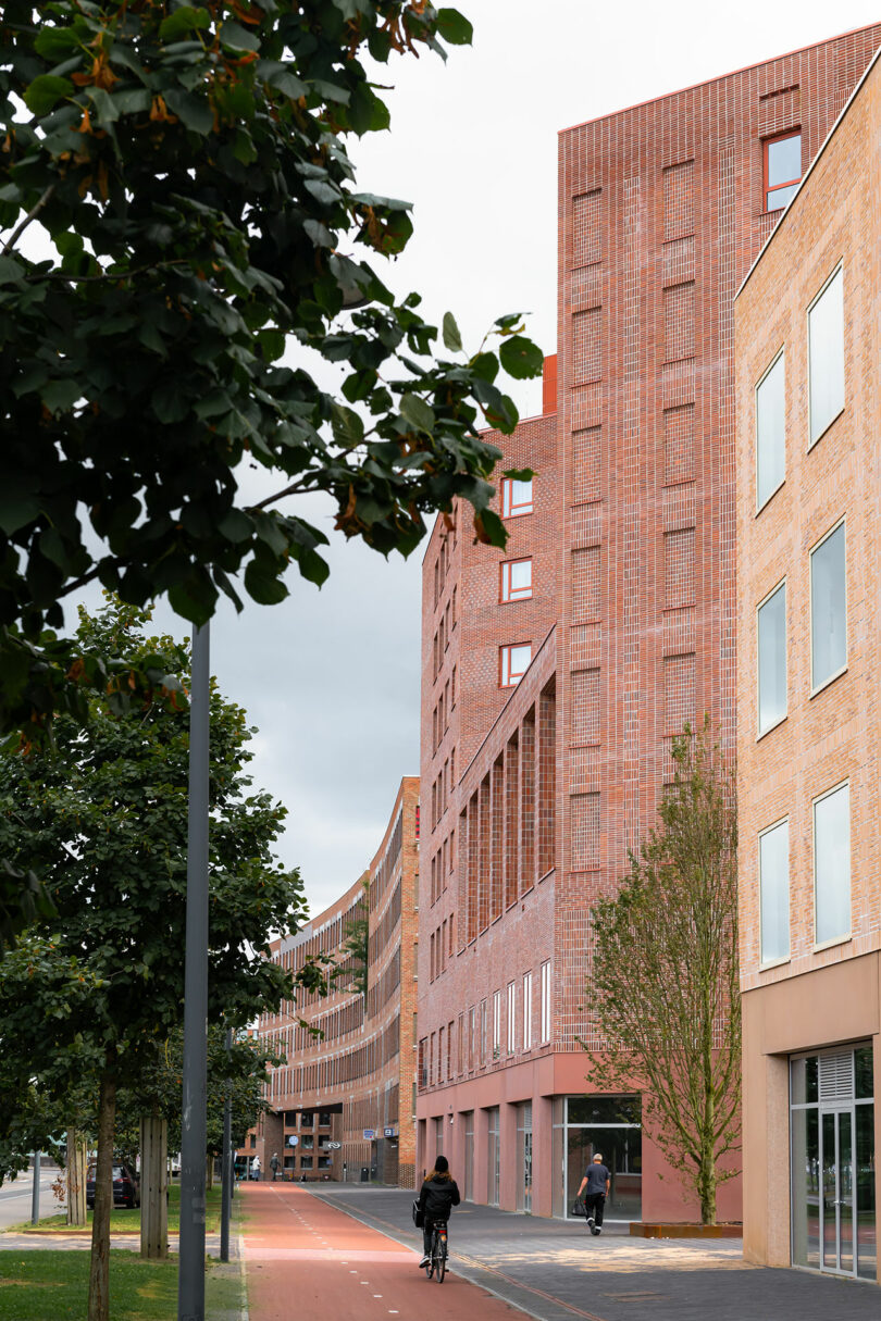 A person rides a bicycle on a red bike lane next to modern brick buildings and trees on a cloudy day.