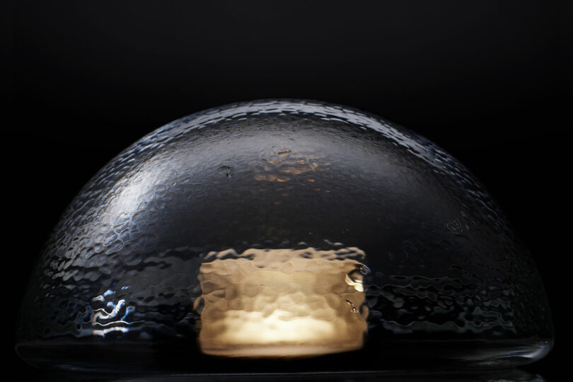 A textured glass dome, reminiscent of the Bocci 14p, covers a lit candle, casting a warm glow on a dark background.