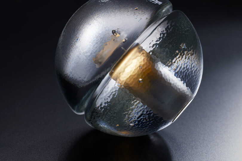 A textured Bocci 14p glass sphere with a metallic cylindrical core rests on a dark surface, illuminated to highlight its reflective and hammered appearance.