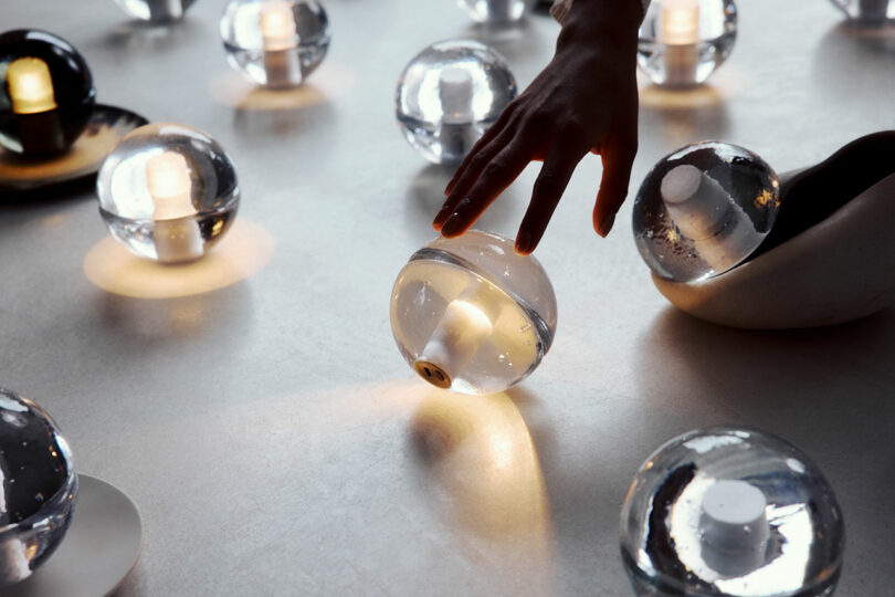 A hand reaches toward a glowing Bocci 14p glass sphere among several similar spheres with internal lights on a smooth, light-colored surface.