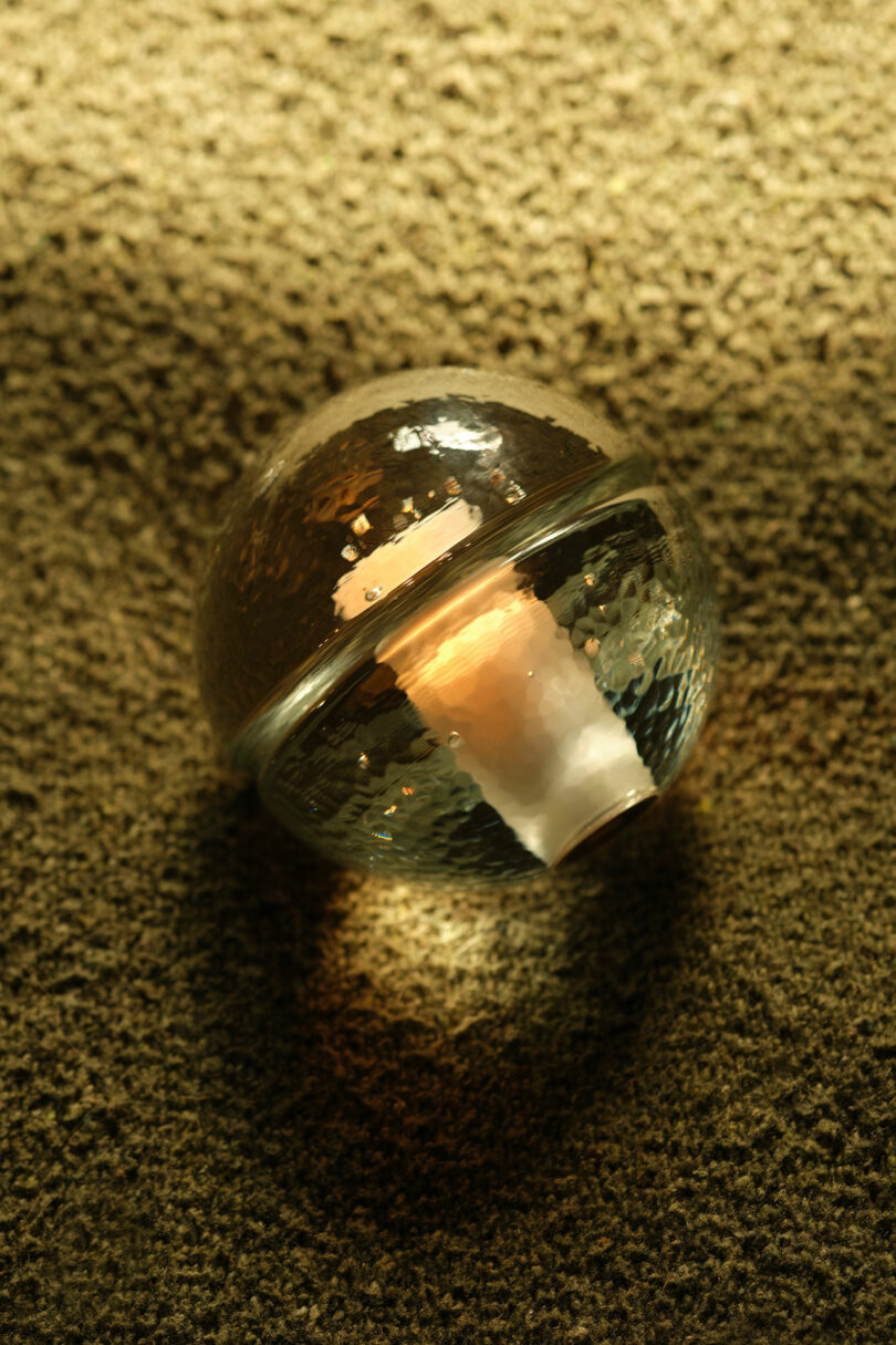 A Bocci 14p glass sphere with a textured surface rests on a carpeted floor, illuminated by warm light.