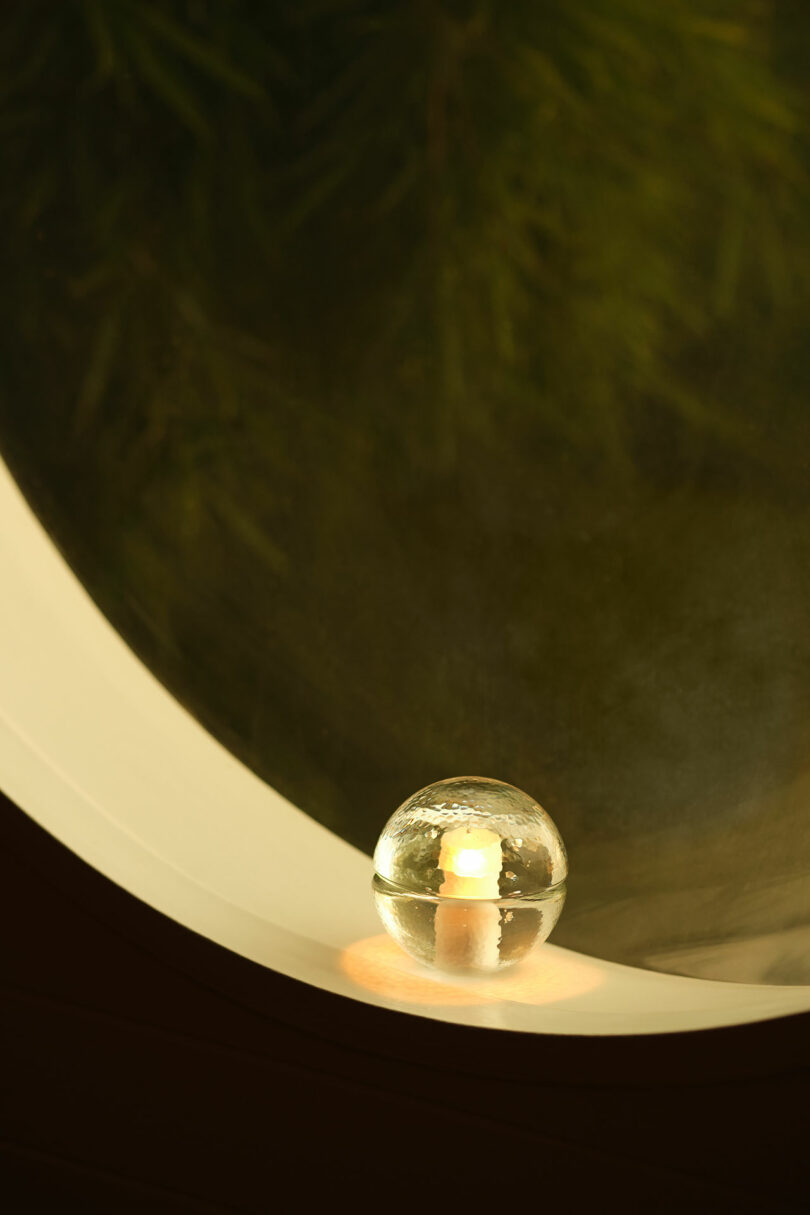 A small Bocci 14p glass sphere with a candle inside sits on a white curved surface, reflecting light, with a blurred green background visible through a circular window.