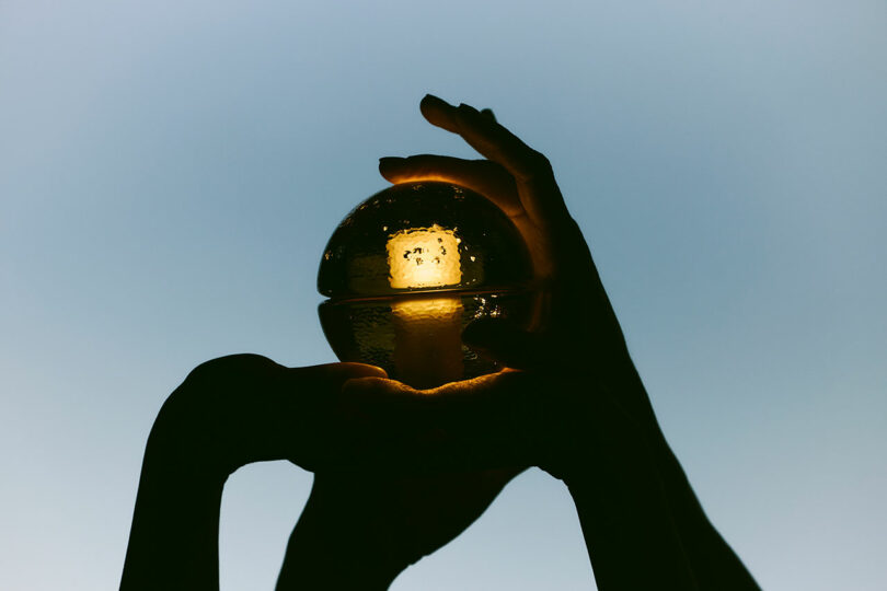 Two hands are holding and framing a glowing Bocci 14p glass sphere against a plain, blue-grey background.