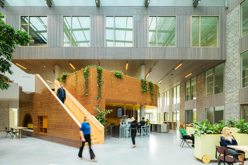 A modern, open atrium with large windows, an orange-tiled staircase, hanging plants, and people walking, sitting, and working in various areas.