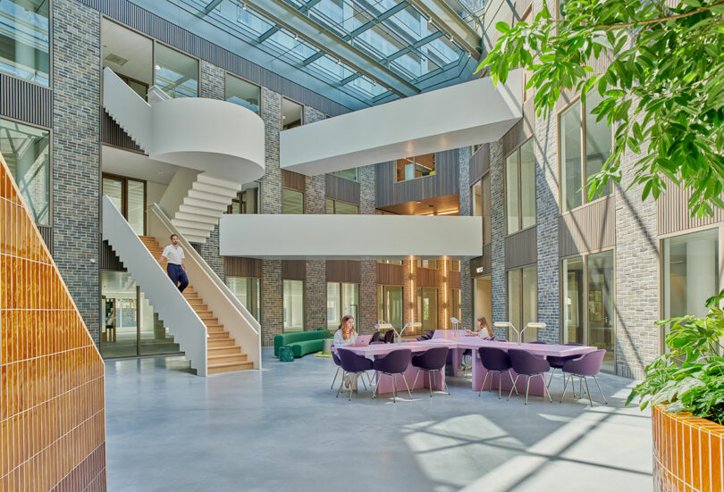 Modern office atrium with a skylight, spiral staircase, people sitting at purple chairs around tables, and greenery in the corner.