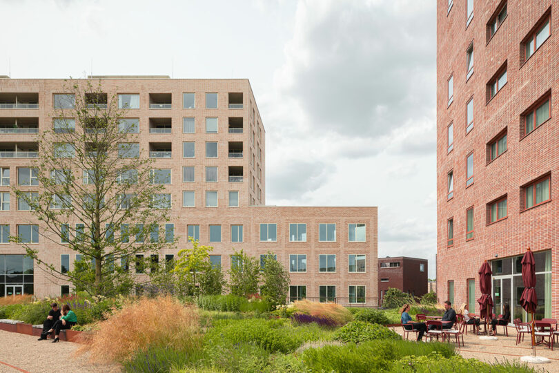 Modern brick apartment buildings surround a landscaped courtyard with greenery, outdoor seating, and people relaxing or conversing on a partly cloudy day.