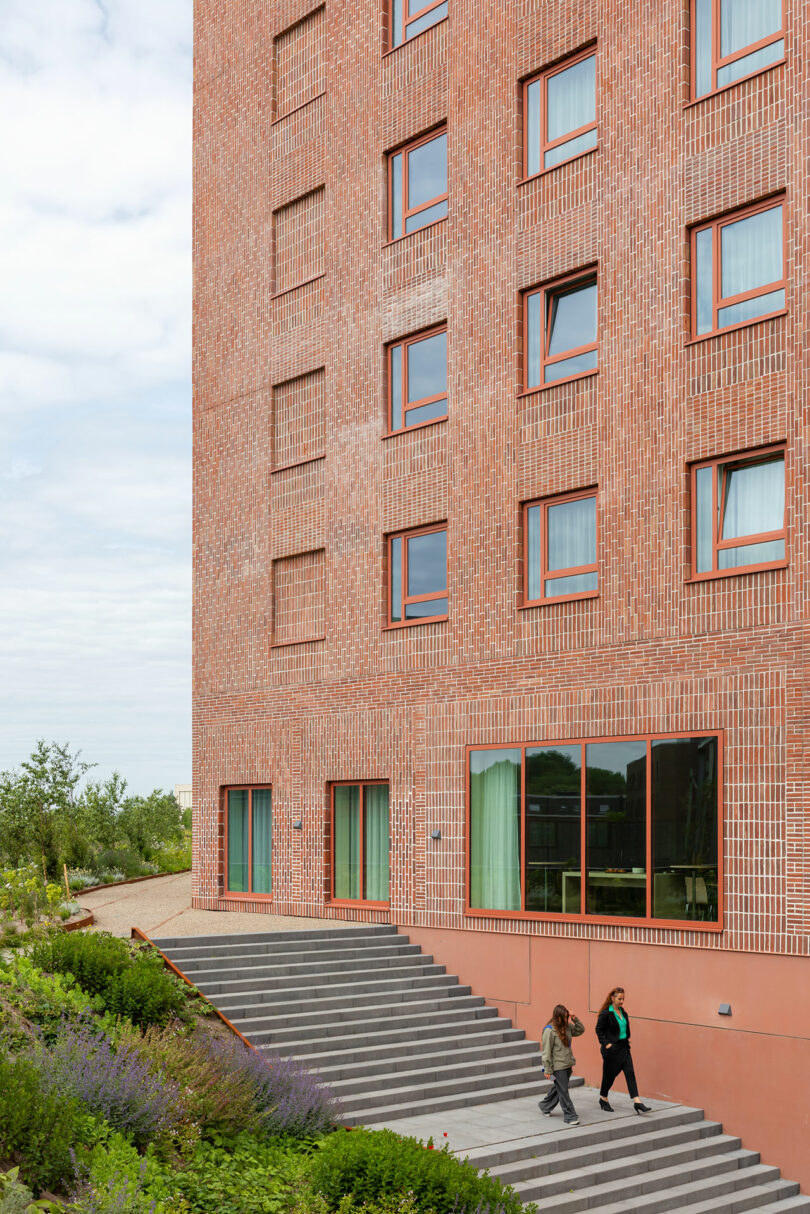 A brick building with large windows, a staircase leading to the entrance, and two people walking down the steps next to landscaped greenery.