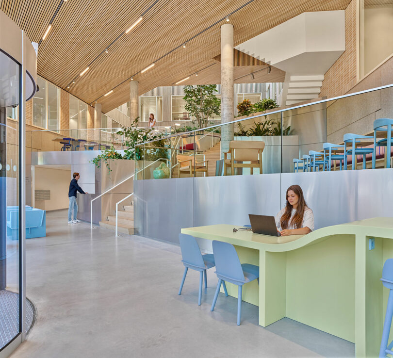 Modern office interior with pastel furniture, glass railings, plants, and people working or walking; large windows and wood ceiling add natural light.