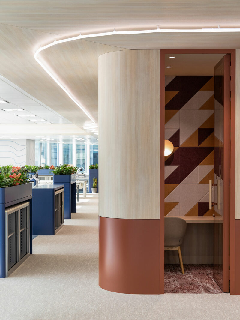 Modern office space with indoor plants, blue desks, and a small private booth featuring geometric wall patterns and a single chair.