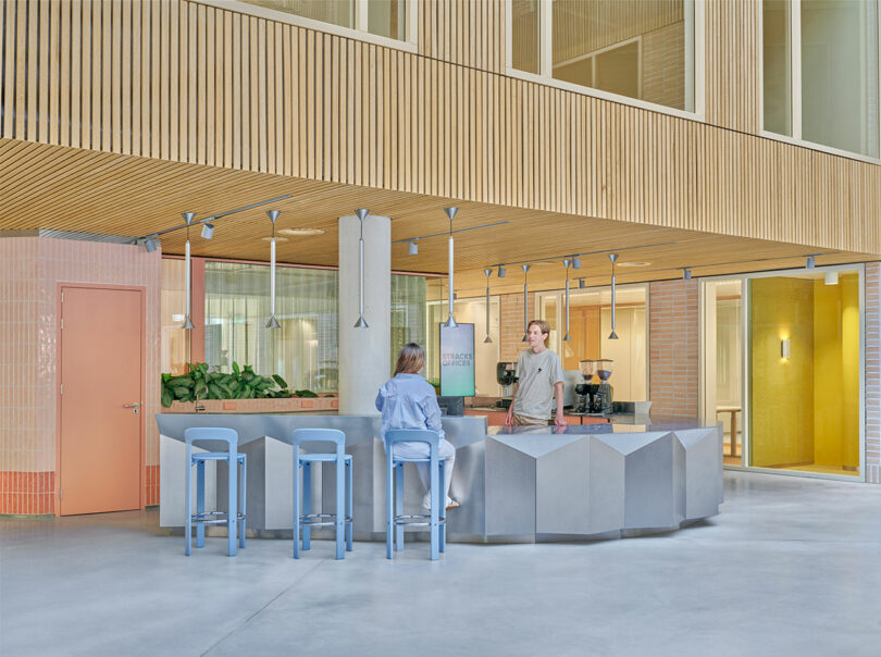 Two people interact at a modern, minimalist café counter with pastel blue stools and wood paneling inside a bright, open space.
