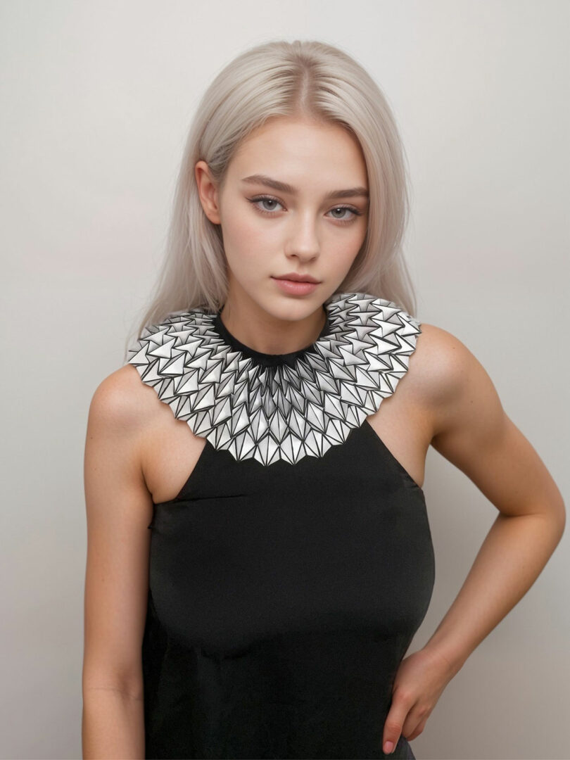 A woman with long, straight platinum blonde hair wears a sleeveless black top and a geometric, ruffled white and black collar from the 3Dtachables Collection. She stands against a plain light background.