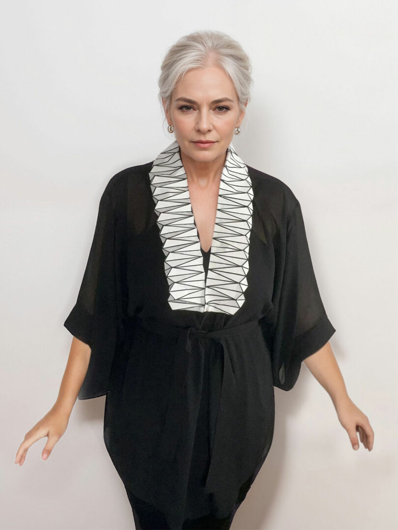 A woman with gray hair in a bun wears a black kimono-style top from the 3Dtachables Collection, featuring a white, geometric-patterned collar, standing against a plain white background.