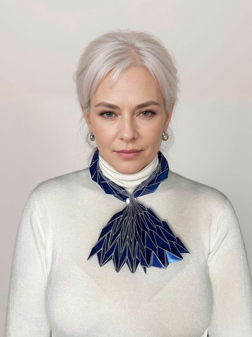 A woman with light hair wearing a white turtleneck sweater and a large geometric blue necklace from the 3Dtachables Collection stands against a plain light background.