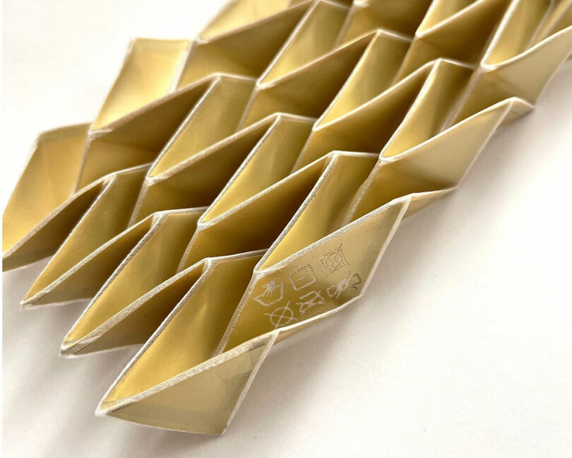 Close-up of gold-colored, diamond-shaped origami pieces from the 3Dtachables Collection, arranged in a repetitive, interlocking pattern on a white background.