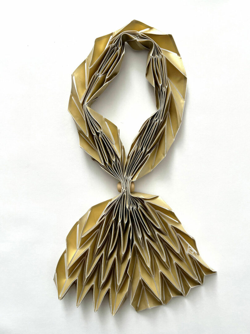 A folded paper sculpture from the 3Dtachables Collection, resembling a scarf, made with gold and white paper arranged in a geometric, pleated pattern and displayed on a white background.