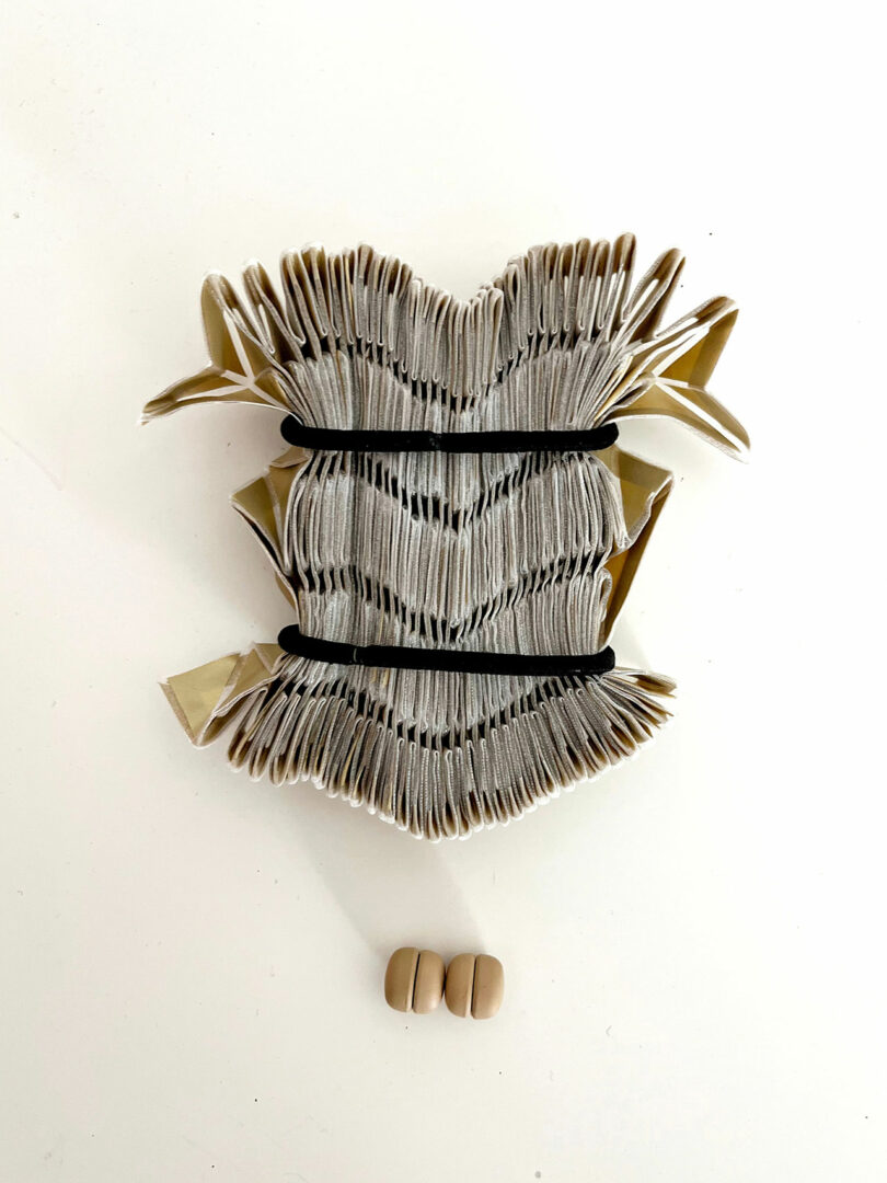 An open, intricately folded book from the 3Dtachables Collection is held by black bands above two pairs of small, round, beige objects on a white surface.