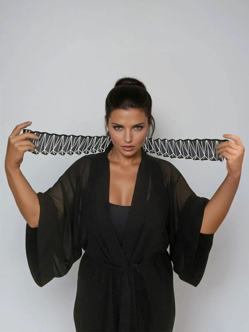 Liat Brandel stands in a black robe, holding a patterned black and white belt stretched in front of her against a plain light background.