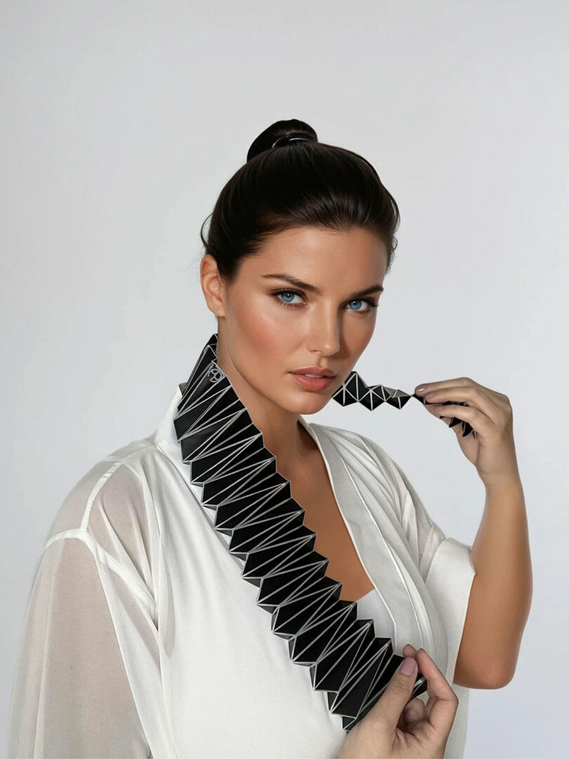 A woman in a white sheer robe, modeled by Liat Brandel, holds a black geometric accordion-like object that wraps around her neck, posing against a plain light background.