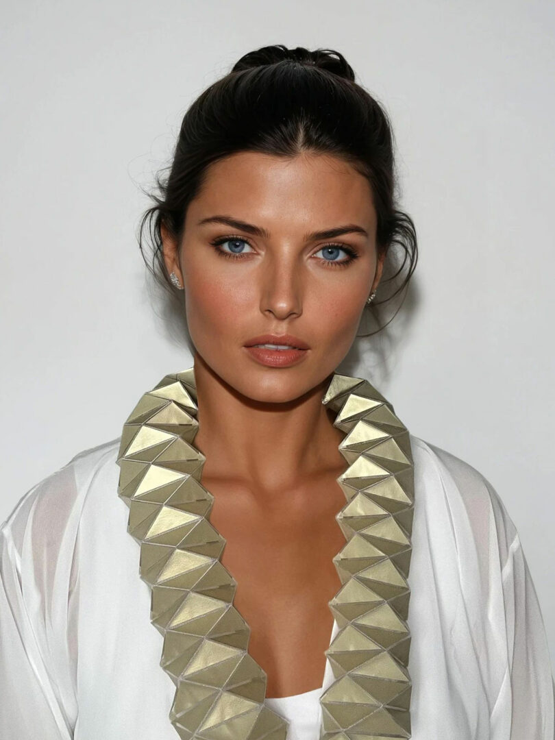 Liat Brandel, a woman with blue eyes and brown hair, wears a white top and a large geometric gold necklace while posing against a plain white background.