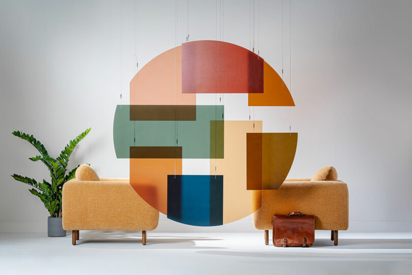 Two yellow sofas face each other with a large, suspended 3form geometric art piece between them; a briefcase sits on the floor and a green potted plant is to the left.