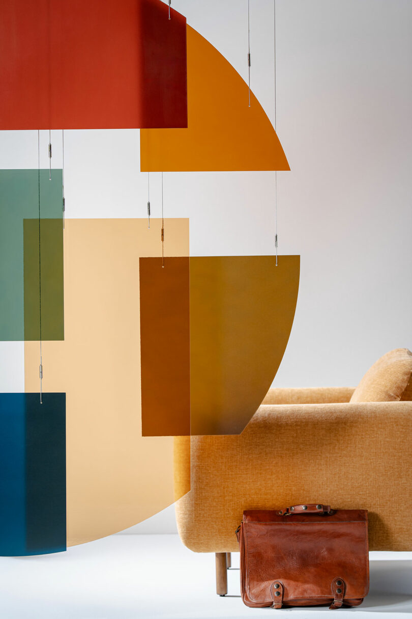 A tan upholstered sofa sits next to a brown leather briefcase; colorful geometric 3form panels hang in the foreground against a plain background.