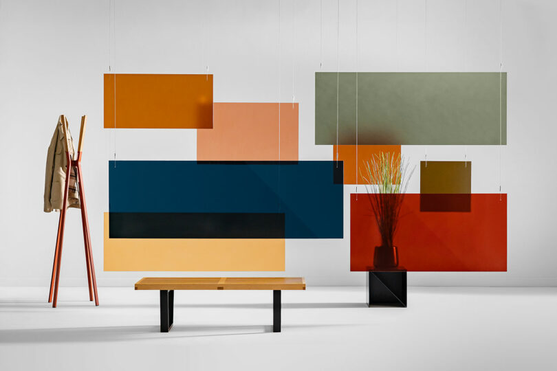 Rectangular 3form colored panels hang from the ceiling in front of a bench, a coat rack with a beige jacket, and a potted plant in this minimalist room.