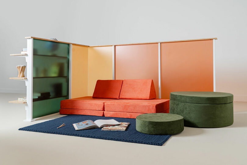 A modern lounge setup with an orange sofa, green ottoman and pouf, blue rug, and a 3form partially frosted glass partition, plus shelving with books against a gradient orange wall.