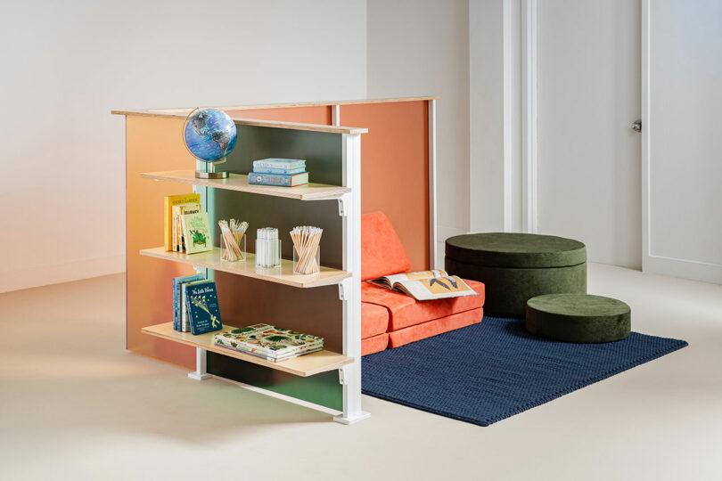 A cozy reading nook features a 3form orange couch, green ottomans, a blue rug, and a bookshelf displaying books, a globe, and cups filled with pencils.