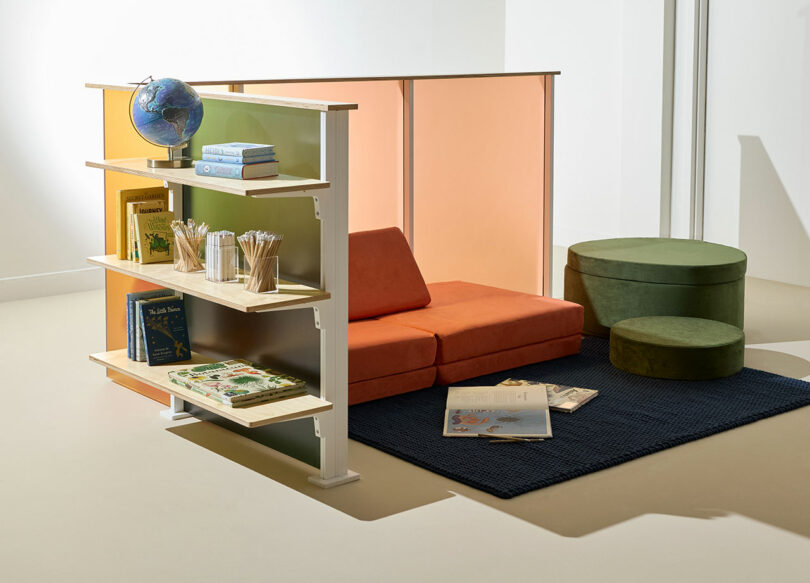 A reading nook with orange seating, green ottomans, a navy rug, a bookshelf holding books and a globe, and a 3form partition in the background.