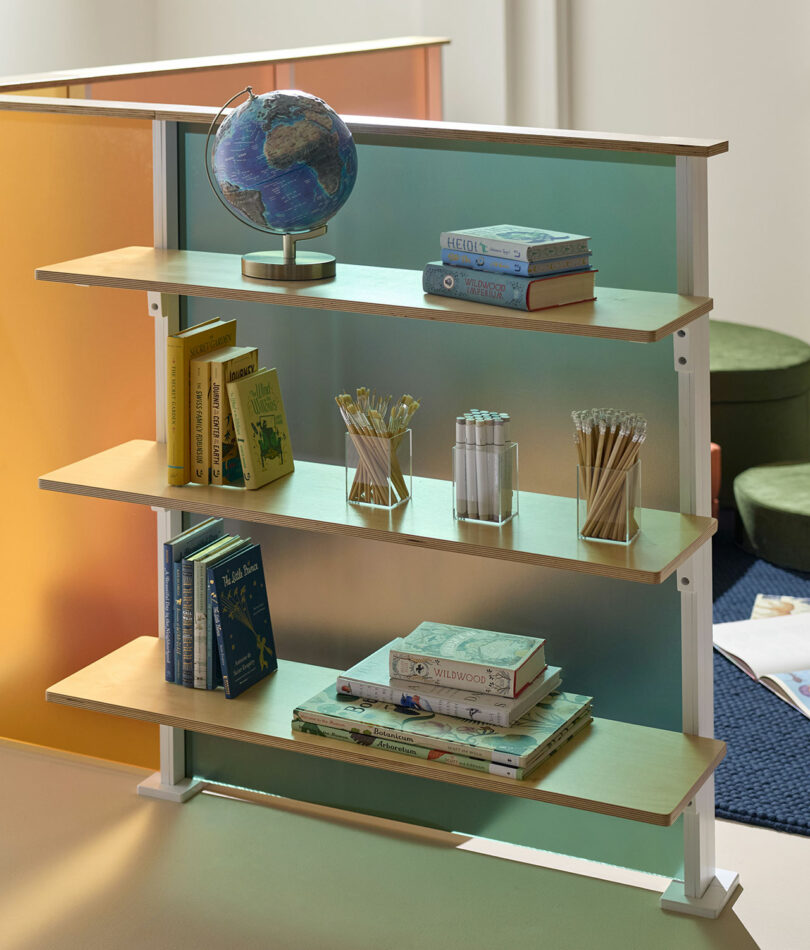 A three-shelf bookcase displaying a globe, books, glass containers with pencils and pens, and a stack of notebooks, set against a colorful 3form partition.