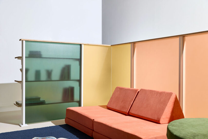 Modern living room with a matte red sofa, pastel yellow and peach partition walls, a green round table, and frosted 3form glass shelving displaying books and decor.