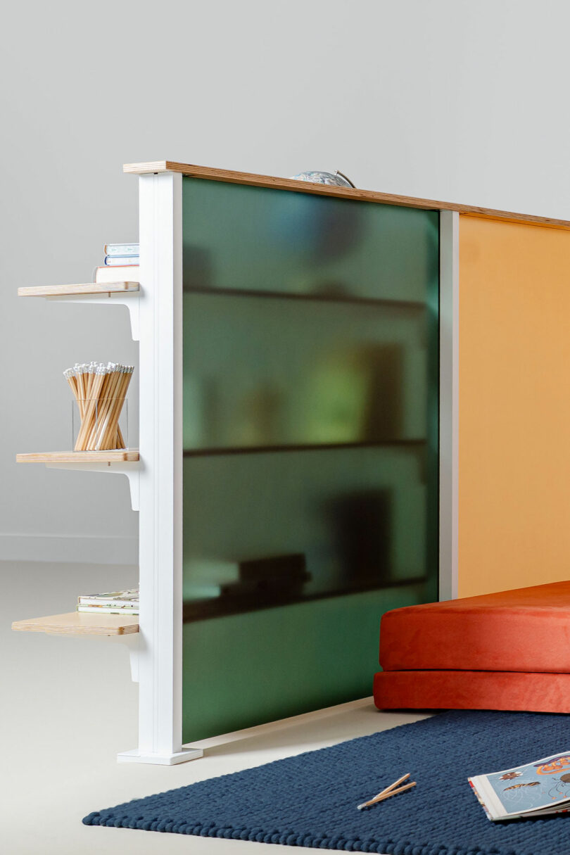 A partial room divider crafted with 3form frosted green glass features integrated wooden shelves holding books and pencils, complemented by an orange cushion and a blue rug on the floor.