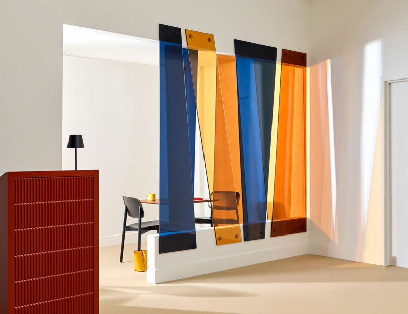 A modern interior featuring colorful 3form translucent panels in blue, orange, and yellow, paired with a black chair and table, a red cabinet, and a minimalist lamp.
