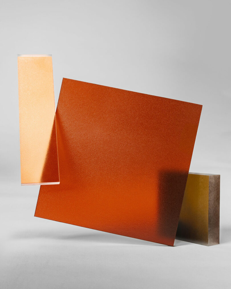 Three rectangular, textured 3form panels—one orange, one beige, and one gold—are arranged abstractly against a plain gray background.
