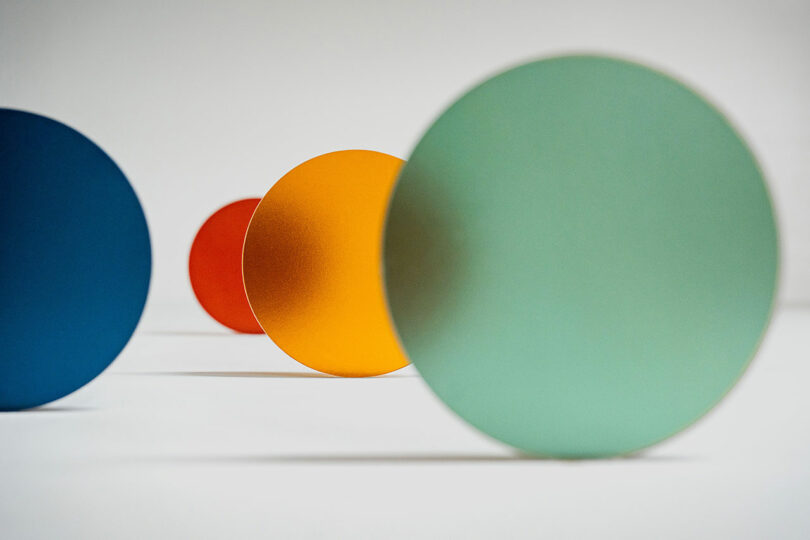 Four colored circular discs—blue, red, orange, and green—stand upright on a white surface in a line, showcasing 3form design innovation with varying focus.