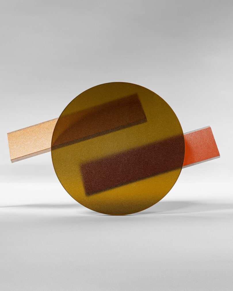 A circular yellow 3form glass sculpture is intersected by two rectangular glass bars, one peach and one orange, set against a neutral background.