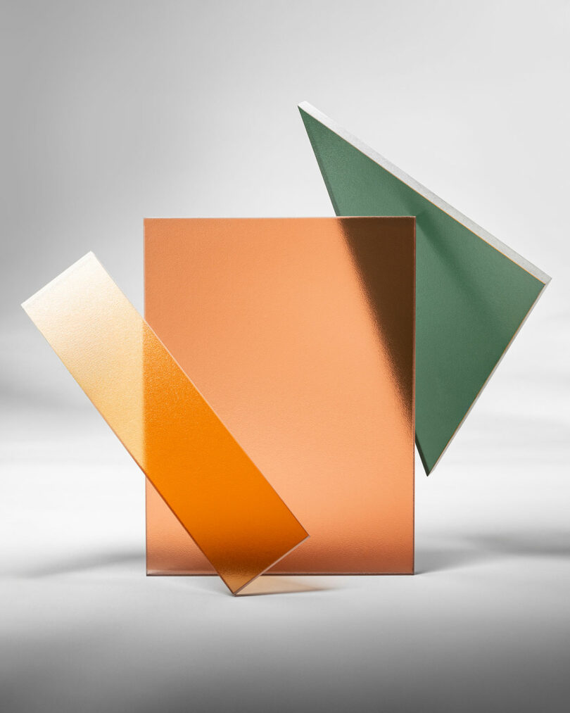 Three geometric glass panels—one orange rectangle, one yellow strip, and one green triangle by 3form—are arranged upright against a neutral background.