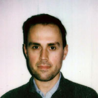 A man with short dark hair and a neutral expression is facing the camera. He is wearing a dark sweater over a collared shirt. The background is plain and light-colored.