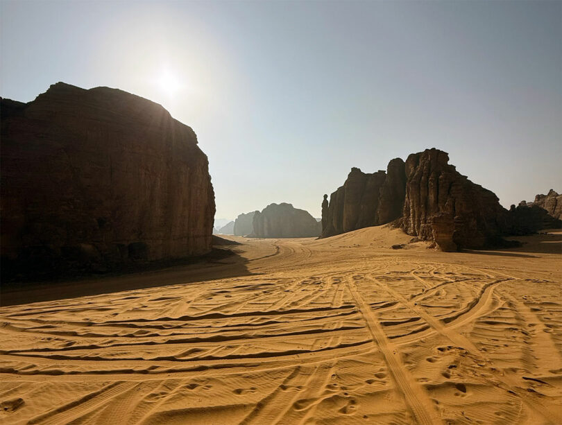 Rock formations and sand dunes with vehicle tire tracks under a bright sky create a stunning AlUla desert landscape.
