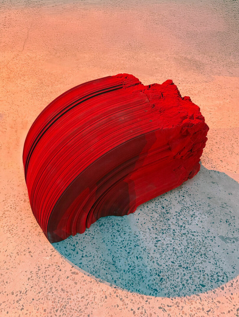 A large, abstract red sculpture with layered, curved lines sits on a speckled concrete floor under warm lighting, evoking the contemporary art scene of AlUla.