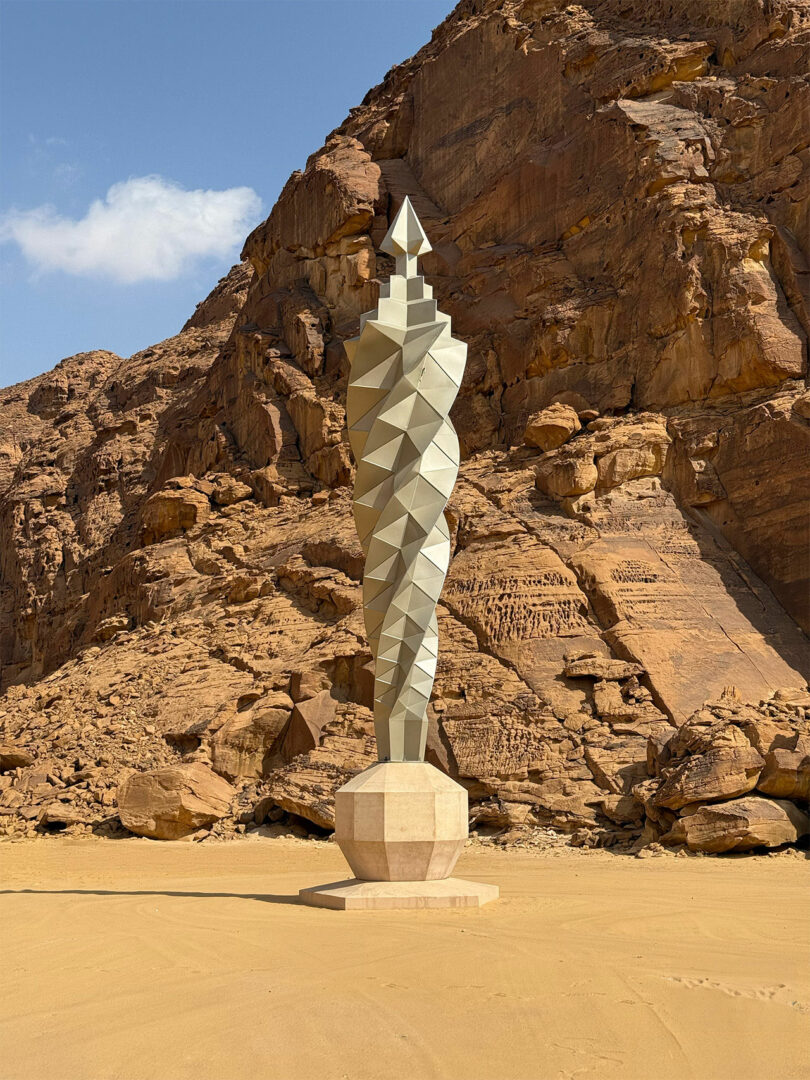 A tall, geometric metal sculpture with a pointed top stands on a sandy base in front of AlUla's rocky desert cliffs under a blue sky.