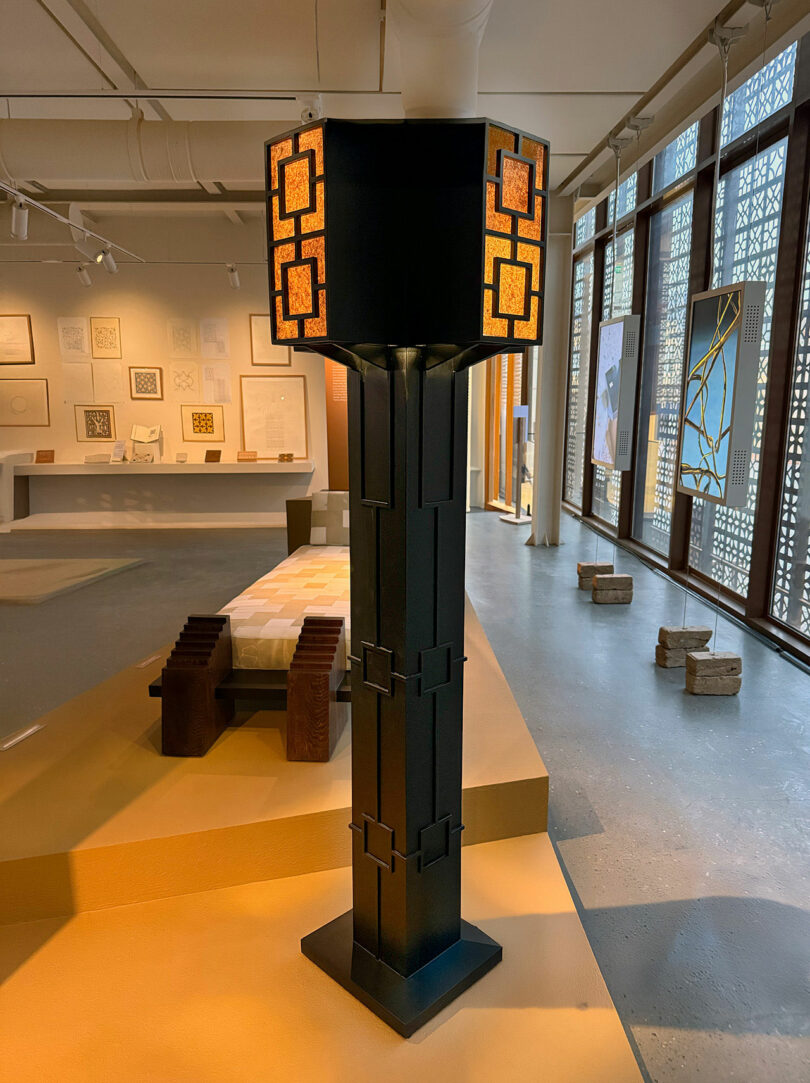 A tall black floor lamp with geometric patterns and amber glass panels stands in a modern, well-lit gallery inspired by AlUla, featuring design displays and patterned windows.