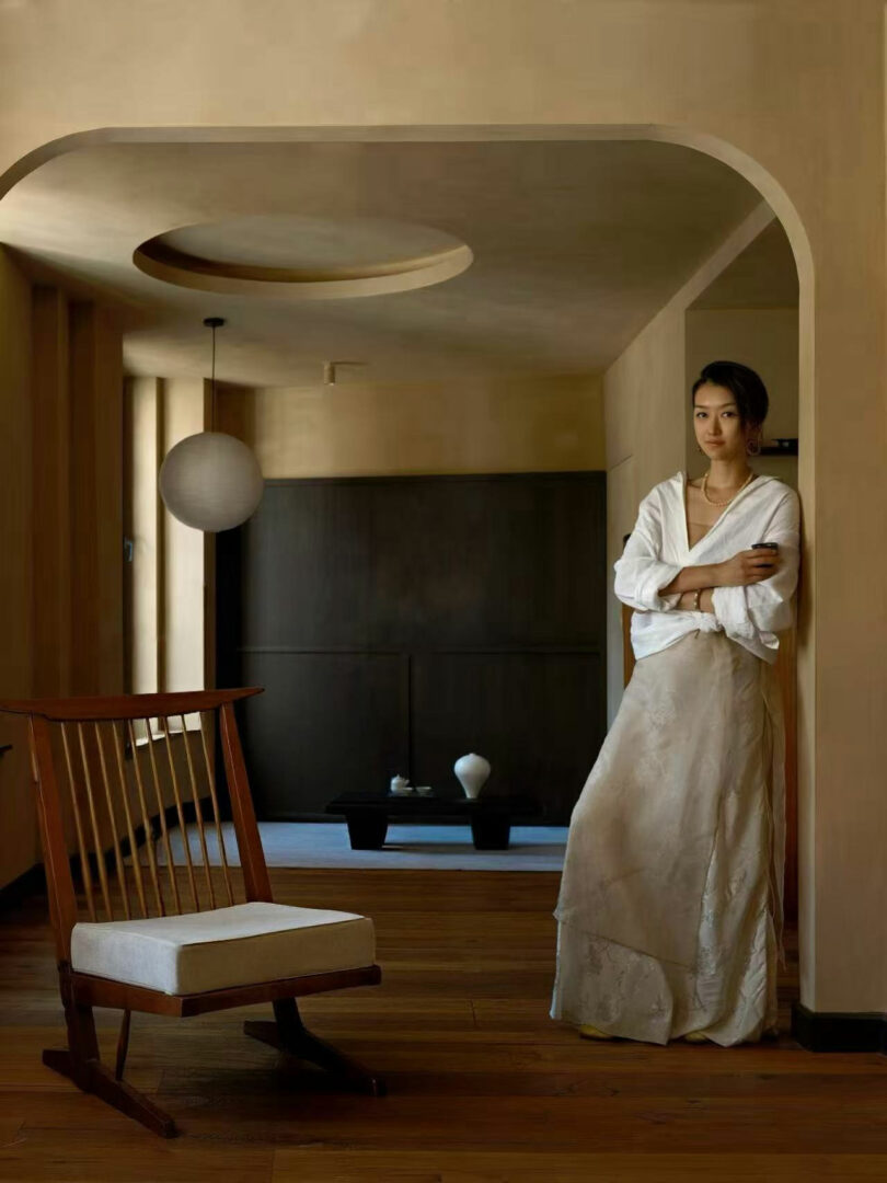 A woman in a white outfit stands against a beige wall in a minimal, warmly lit room with wooden floors, a wooden chair, an Amber Pan on display, and a round hanging light.