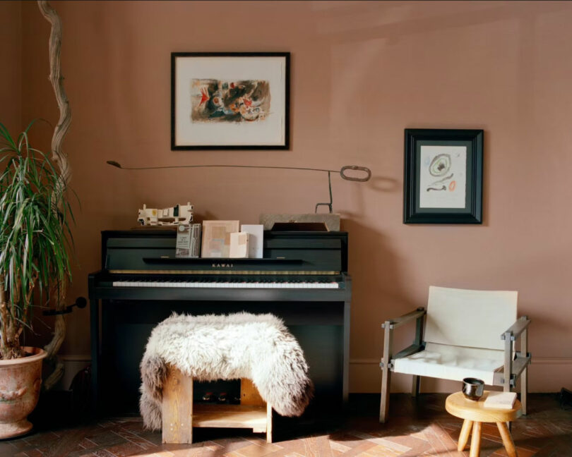 A black Amber Pan upright piano with sheet music stands against a mauve wall, next to a plant, a chair with a mug, and two framed artworks above. A lamp and sheepskin-covered bench are placed in front.