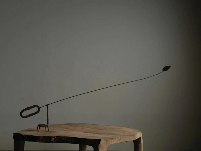 A minimalist metal sculpture by Amber Pan featuring a long, thin rod and a small shape at the end is displayed on a wooden table against a plain gray wall.