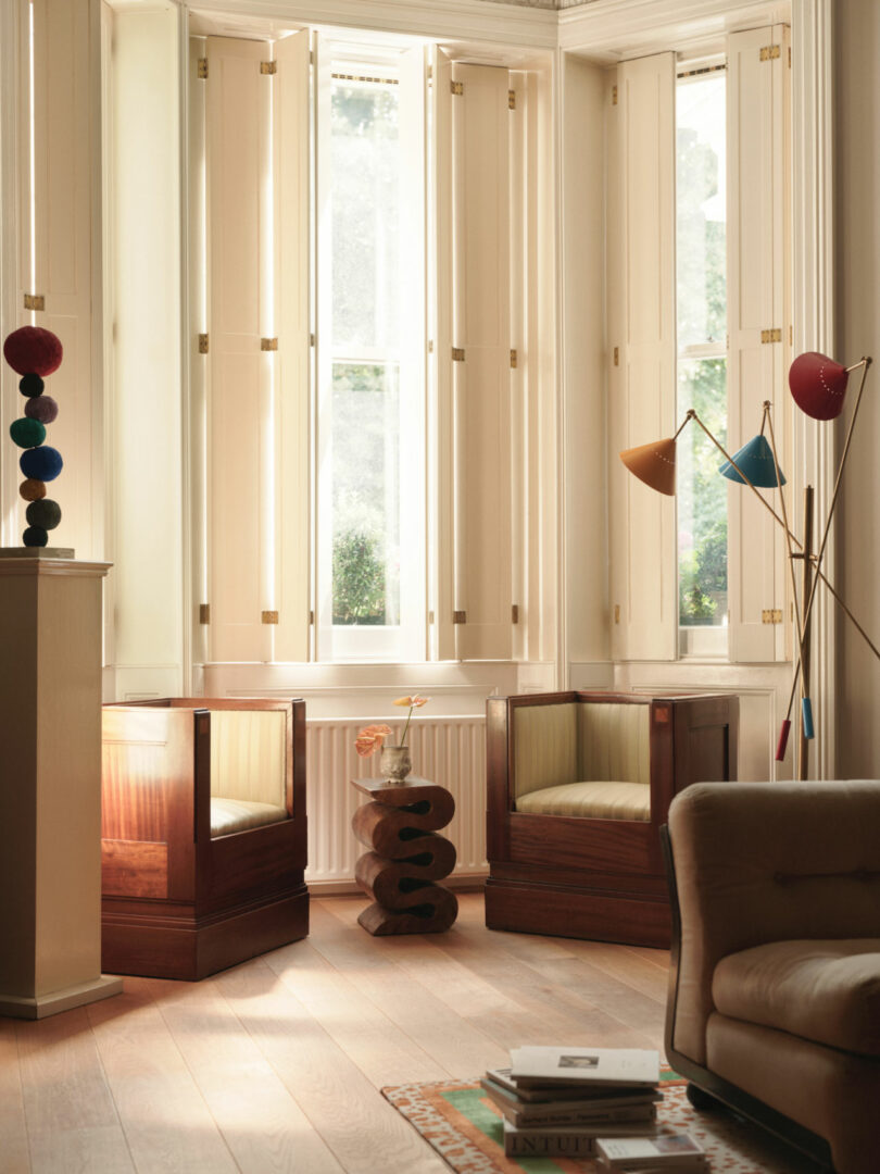A sunlit sitting area with two wooden armchairs, a sculptural side table, modern lamps, and tall windows with cream shutters is softly illuminated by an amber pan of light.