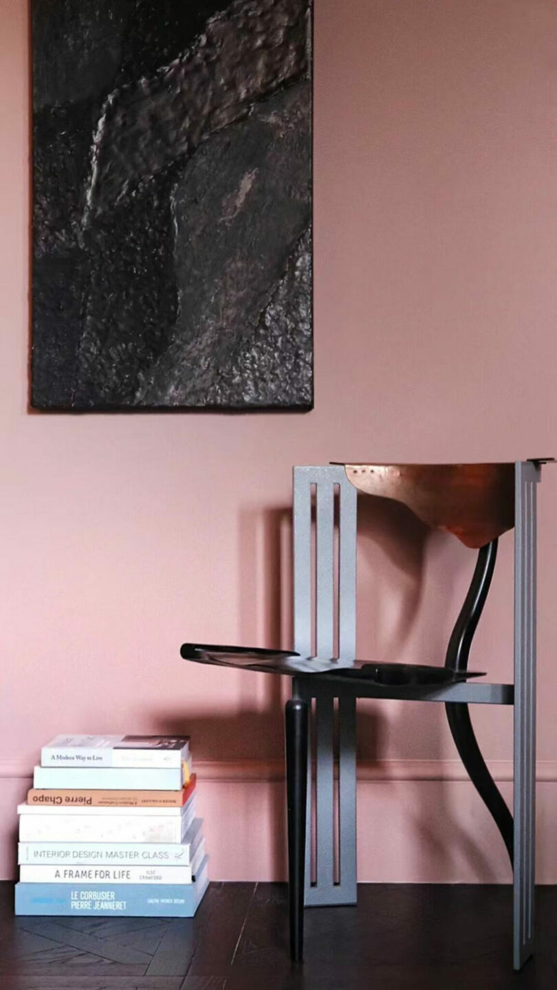 A modern chair is placed against a pink wall with a textured black artwork by Amber Pan above it. A stack of design and art books sits on the dark wooden floor to the left of the chair.