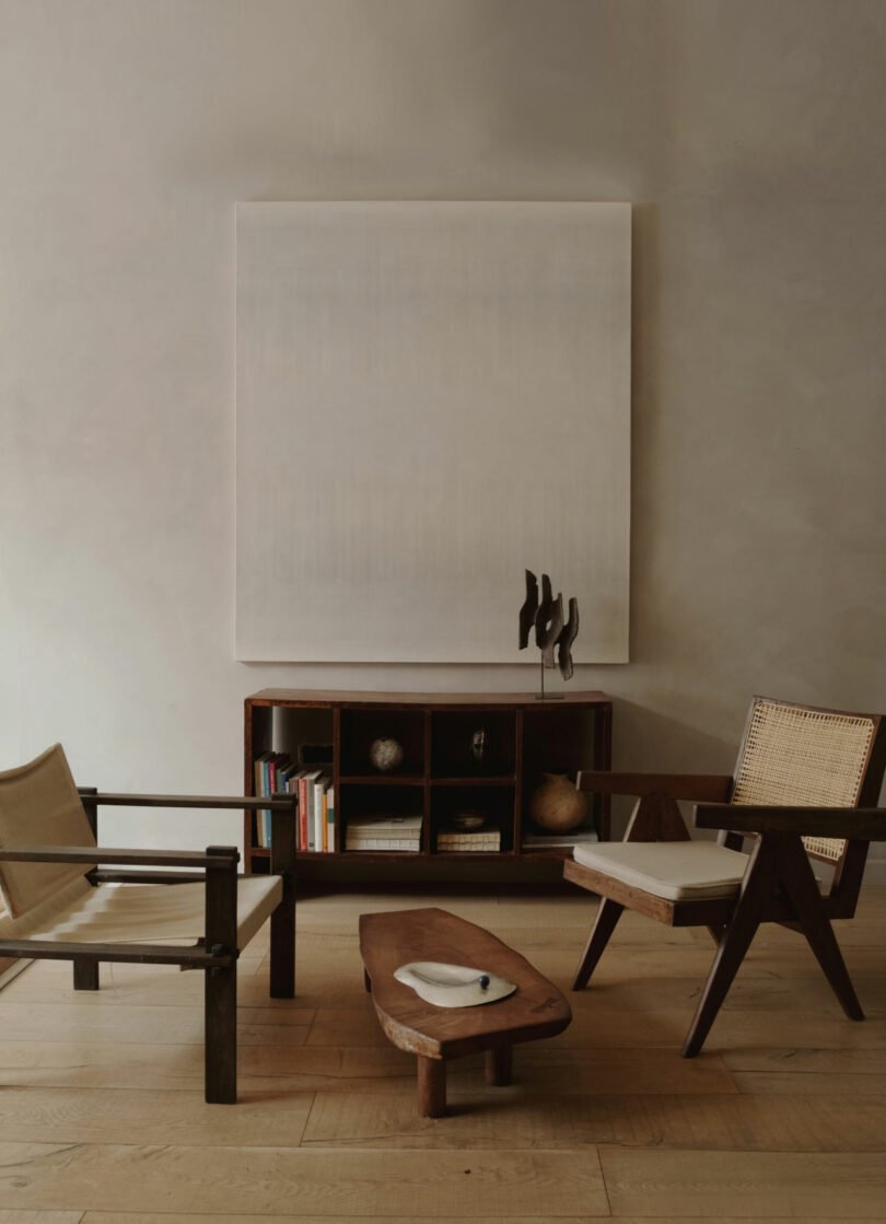 A minimalist living room with two wooden chairs, a low wooden coffee table, a shelf with books and decor, and a large abstract painting featuring warm amber pan tones on the wall.