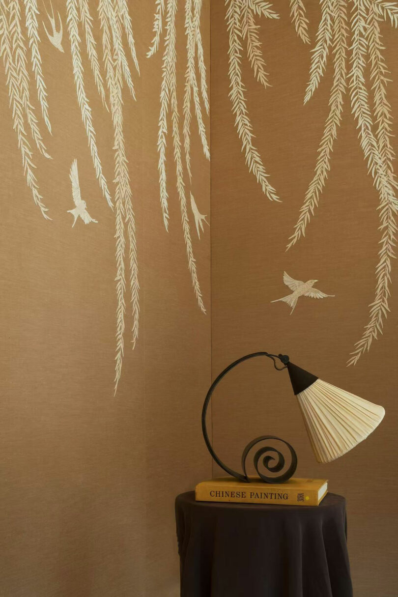A lamp with a spiral base by Amber Pan sits on two books, one titled "CHINESE PAINTING," against a wall decorated with white bird and willow branch designs.