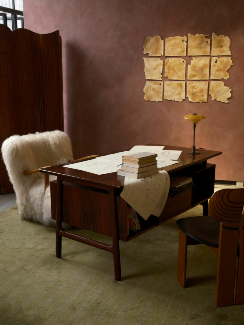 A mid-century wooden desk with scattered papers and stacked books is paired with a fuzzy white chair; an Amber Pan rests nearby as abstract artwork hangs on a mauve wall in a softly lit room.